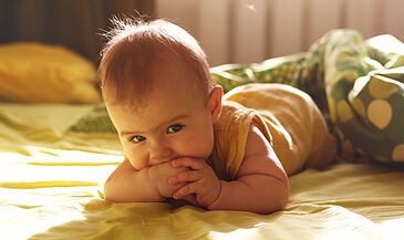 A baby with light brown hair lies on a bed, smiling and chewing on a hand. The room is softly lit with yellow and green bedding.